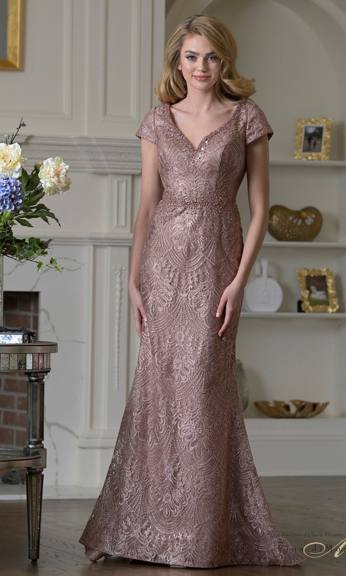 A woman stands indoors in the Rina Di Montella RD3532, a dark mauve floor-length formal dress with lace beading, short sleeves, V-neckline, and mermaid silhouette—ideal for a fashionable mother-of-the-bride.